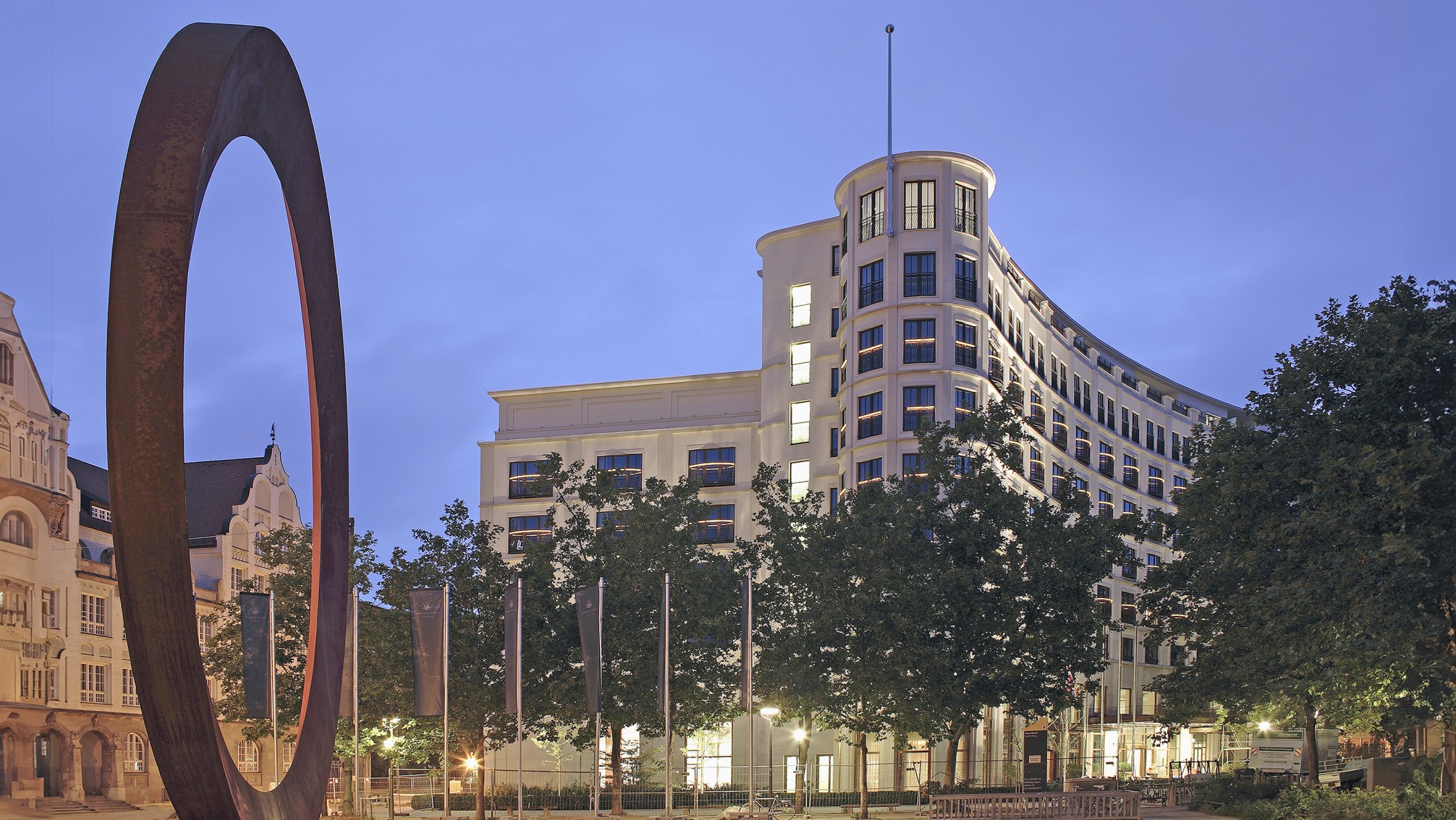 Nachts beleuchtete Fassade des Charles Hotels mit Fahnen und Kunstwerk im Vordergrund in den Lenbach Gärten in München